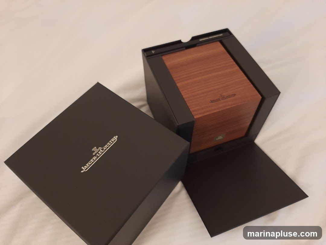 A dark gray box with the Jaeger-LeCoultre logo in silver is open, revealing a wooden inner box. The wooden box also has the Jaeger-LeCoultre logo.
