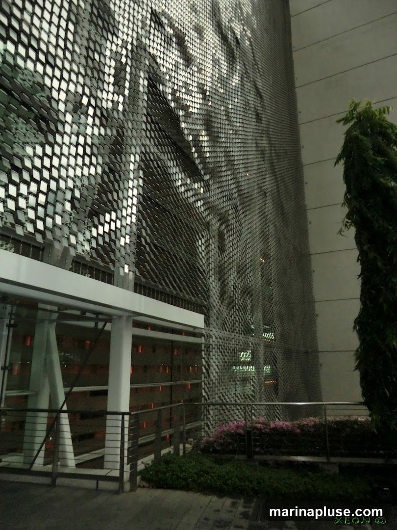 A close-up view of a building's exterior, featuring a unique, textured facade composed of numerous small, reflective squares arranged in a diamond pattern. The facade creates a dynamic visual effect, with light reflecting off the surfaces and casting shadows. To the left, a modern entrance structure with glass and metal railings is visible. In the foreground, there is a planter with pink flowers and green foliage, and a tree stands to the right. The overall impression is one of contemporary architecture.