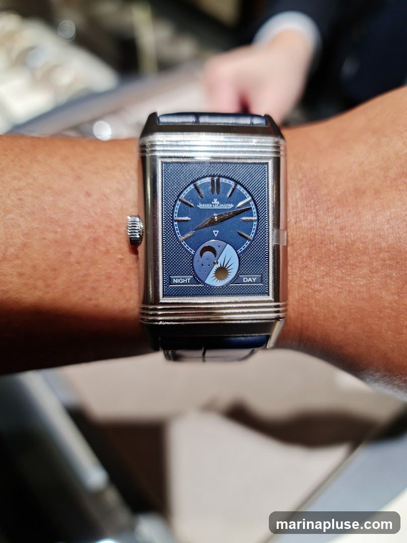A close-up shot of a Jaeger-LeCoultre watch on a person's wrist. The watch has a rectangular silver case and a dark blue dial with a day/night indicator. The brand name "JAEGER-LeCOULTRE" is visible on the dial.