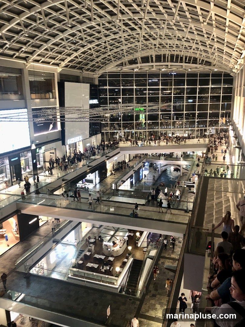An aerial view of a multi-level shopping mall with a large glass ceiling and numerous shops and people. There are escalators, walkways, and a central atrium with a water feature. Several branded storefronts are visible, including Chanel and Gucci.