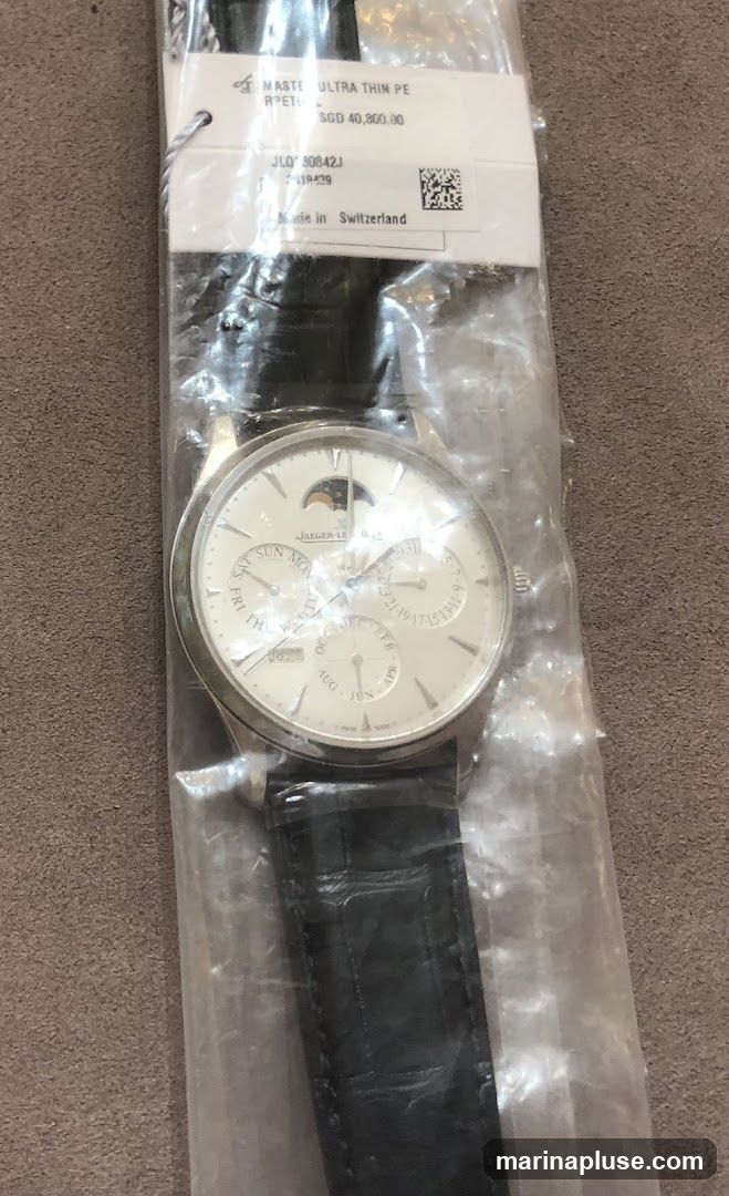 A close-up of a Jaeger-LeCoultre Master Ultra Thin Perpetual watch, still in its plastic packaging. The watch has a white dial with a moon phase complication at the top. Sub-dials display the day of the week, date, and month. The watch is fitted with a black leather strap. A tag attached to the packaging shows the model name, price (SGD 40,800.00), reference number, and indicates it is made in Switzerland.