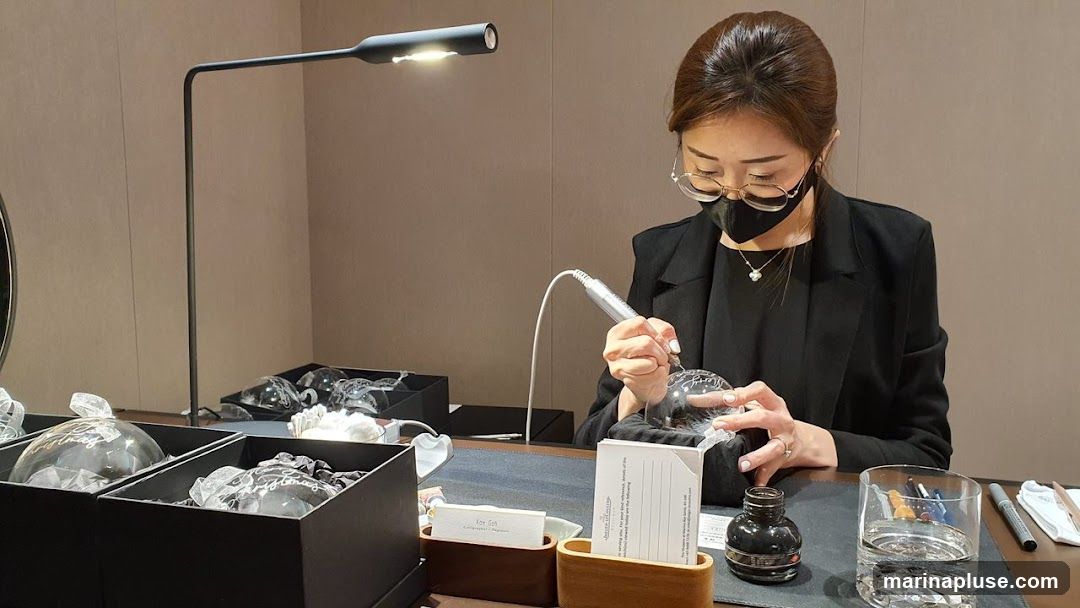 A woman wearing glasses and a mask is engraving a glass ornament with a tool. She is seated at a table with several other ornaments in boxes, a lamp, an inkwell, and a glass of water.