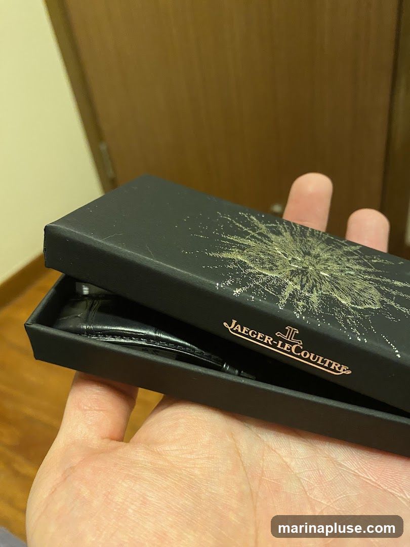 A black box with a gold abstract design and the Jaeger-LeCoultre logo is open, revealing a black watch with a textured strap inside. The box is held by a person's hands.