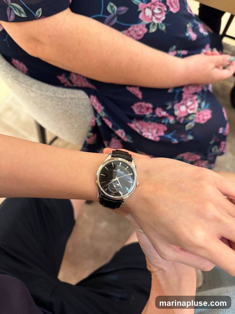 A close-up shot of a person's wrist wearing a black-faced watch with a leather strap. The watch has a silver case and a dial with hour markers and a moon phase complication. The person is wearing dark clothing. Another person in a floral dress is visible in the background.