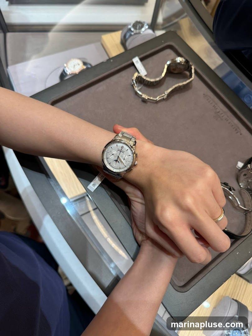 A person's arm is shown wearing a silver watch with a white face and multiple subdials. The watch is on a display tray with other watches.