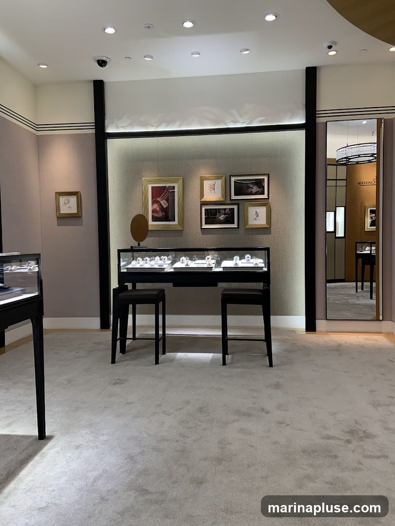 A well-lit jewelry store interior with display cases and artwork. In the center, a long display case with jewelry is flanked by two dark stools. Above the case, a collection of framed pictures adorns the wall. To the right, a large mirror reflects another section of the store, including a chandelier and display cases. The floor is covered in a light-colored carpet.