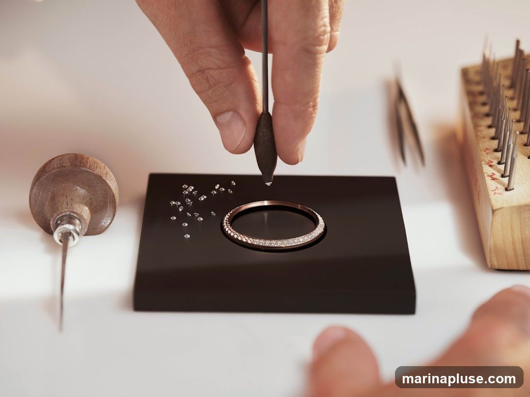 A close-up shot shows a jeweler's hands meticulously setting small diamonds onto a rose gold ring. Various tools, including a graver, tweezers, and a wooden tool holder with pointed instruments, are visible on the white surface, suggesting a workshop environment.