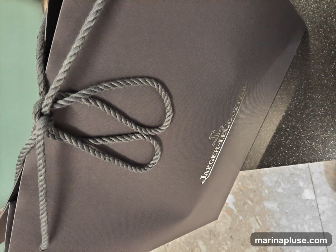 A gray shopping bag with a rope handle tied in a knot. The Jaeger-LeCoultre logo is embossed on the side of the bag.