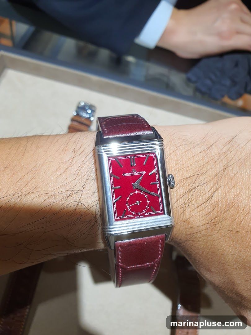 A close-up shot of a rectangular watch with a red face and a burgundy leather strap on a person's wrist. The watch has a small seconds subdial. The brand name "JAEGER-LECOULTRE" is visible on the watch face. In the background, another watch and a person's hands in a suit jacket are visible.
