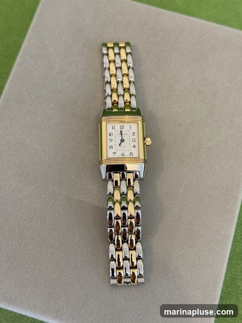 A rectangular watch with a gold-tone case and a two-tone stainless steel bracelet is displayed on a gray surface. The watch face is white with black Arabic numerals and blue hands. The brand name "Jaeger-LeCoultre" is visible on the dial.