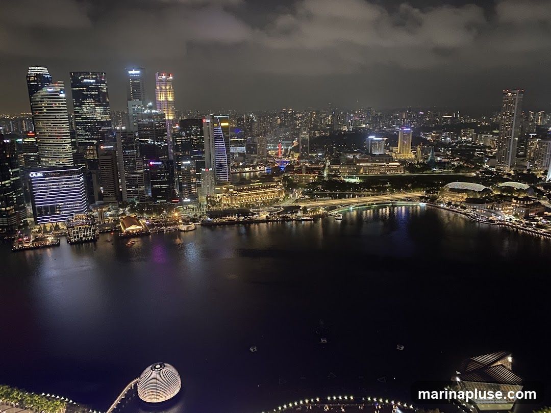 A panoramic view of the Singapore skyline at night, showcasing illuminated skyscrapers and the Marina Bay area. The water reflects the city lights, creating a vibrant and dynamic scene.