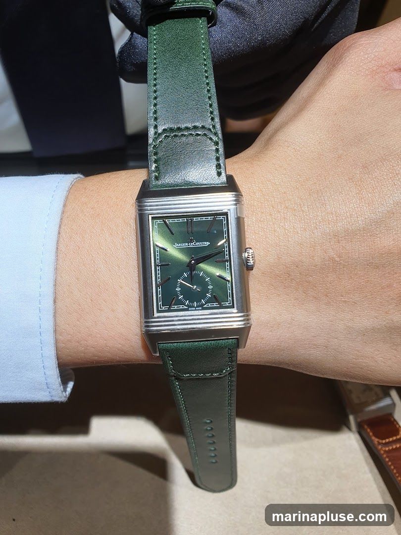 A close-up shot of a person wearing a rectangular Jaeger-LeCoultre watch with a green dial and a matching green leather strap. The watch face features silver hour markers and hands, with a small seconds subdial at the 6 o'clock position. The watch is being held by a gloved hand, suggesting it is on display or being presented.