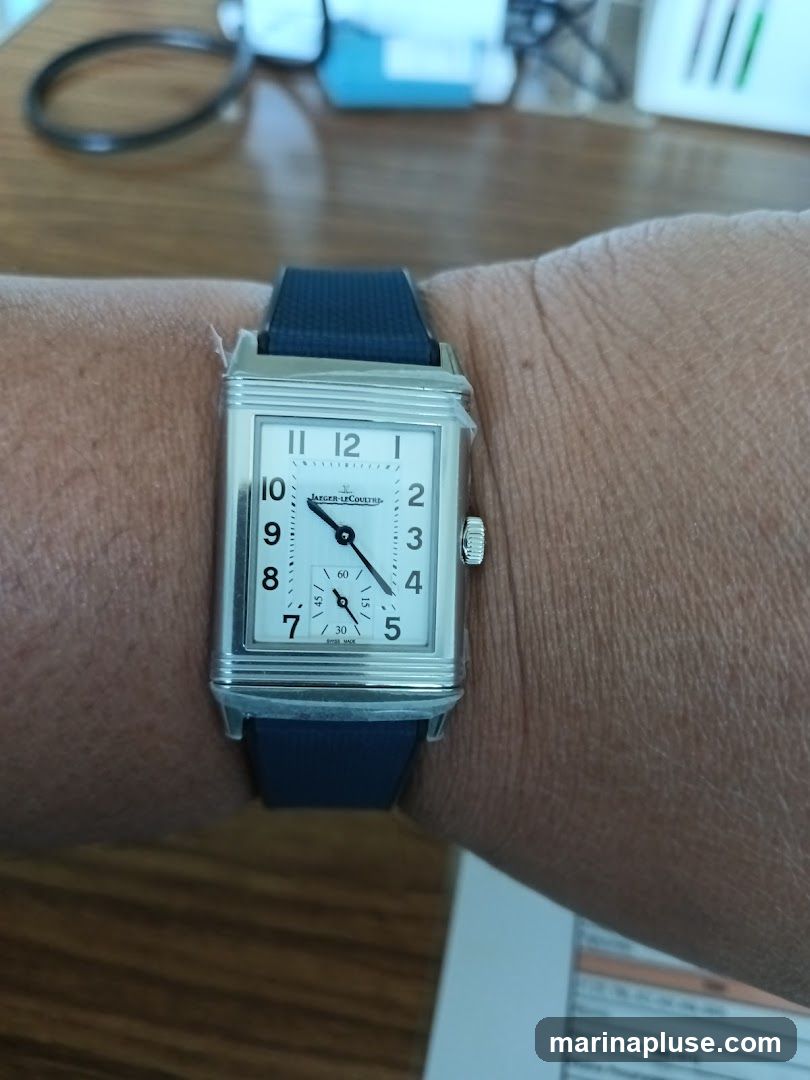 A close-up shot of a rectangular Jaeger-LeCoultre watch with a white dial and black Arabic numerals on a person's wrist. The watch has a dark blue strap. The background shows a wooden surface and some out-of-focus items.