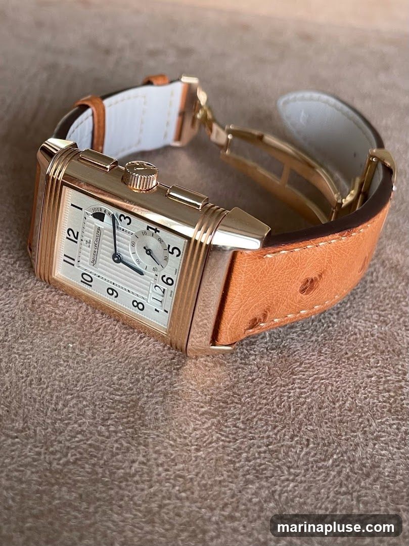 A close-up shot of a rectangular Jaeger-LeCoultre watch with a rose gold case and a white dial. The watch has Arabic numerals for hours, a small seconds subdial, and a date window. It is fitted with a tan ostrich leather strap with white stitching and a rose gold deployant clasp. The watch is resting on a textured brown surface.