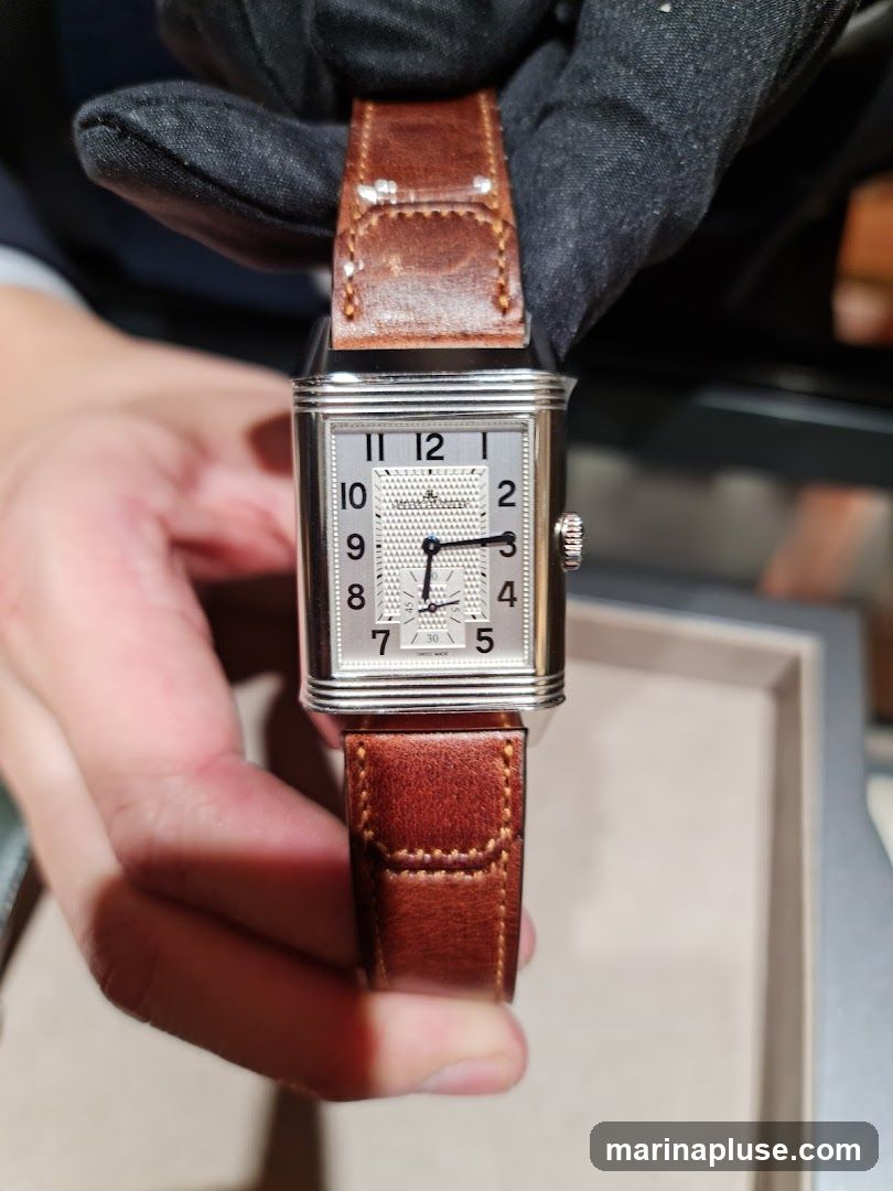 A close-up shot of a rectangular Jaeger-LeCoultre watch with a silver dial and brown leather strap. The watch is held by hands wearing black gloves and a white shirt sleeve. The dial features Arabic numerals for hours and a smaller sub-dial. The brand name "JAEGER-LECOULTRE" is visible at the top of the dial.