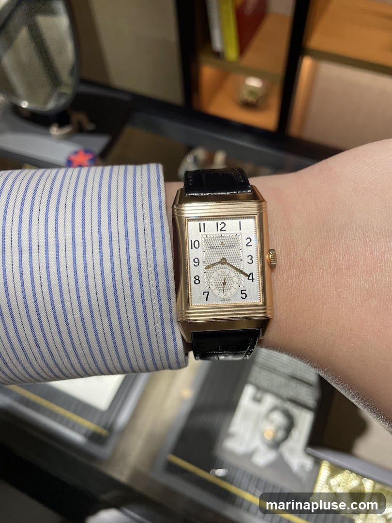 A close-up shot of a person's wrist wearing a rectangular gold watch with a black leather strap. The watch face is white with black Arabic numerals and a small sub-dial. The person is wearing a blue and white striped shirt. The background appears to be a display case in a store, with blurred items and a black and white photograph visible.
