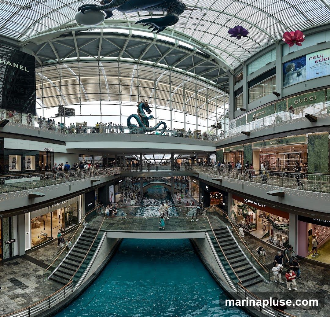 A panoramic view of the interior of a multi-level shopping mall. A bright blue canal runs through the center of the mall, with bridges and walkways crossing over it. Several escalators and staircases provide access to different levels. Shops with prominent signage, including CHANEL and GUCCI, are visible on various floors. Large decorative dragon sculptures and floral-like installations hang from the ceiling, which is a glass dome. Many shoppers are seen browsing and walking around the mall.