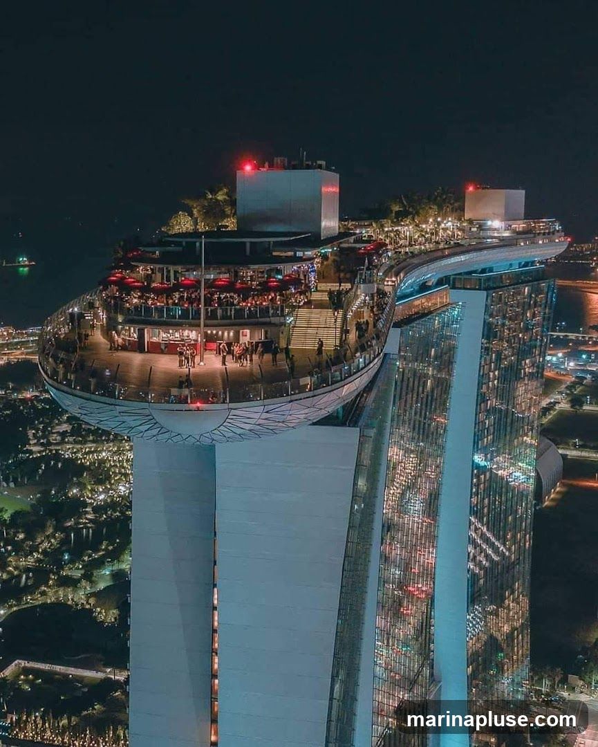 An aerial view at night of a rooftop bar and restaurant situated atop a modern skyscraper. The establishment features an expansive outdoor seating area with numerous tables, many covered by red umbrellas. People are visible mingling and dining on the deck, which is illuminated by warm lighting. The bar area is also visible, with patrons gathered around. The skyscraper itself is a prominent landmark, with its glass facade reflecting the city lights below. The surrounding cityscape is visible in the background, with lights twinkling across the urban landscape.