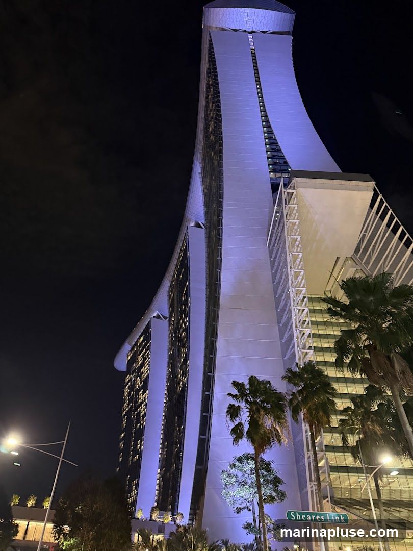 A low-angle night shot of the Marina Bay Sands hotel in Singapore. The iconic building is illuminated with purple lights, highlighting its distinctive architecture. Palm trees are visible in the foreground, and a street sign for 'Sheares Link' is present. The sky is dark and cloudy.
