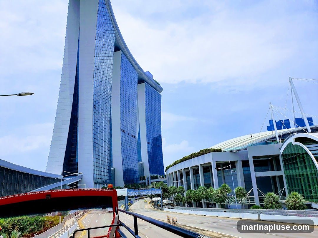 A wide shot of the Marina Bay Sands complex in Singapore under a bright, cloudy sky. The iconic three-tower hotel, with its distinctive boat-shaped top, dominates the left side of the frame. To the right, a large, modern building with a curved, white roof and glass facade is visible. A road with traffic runs in the foreground, and lush greenery surrounds the structures. A red bus with text on its side is partially visible in the lower left.