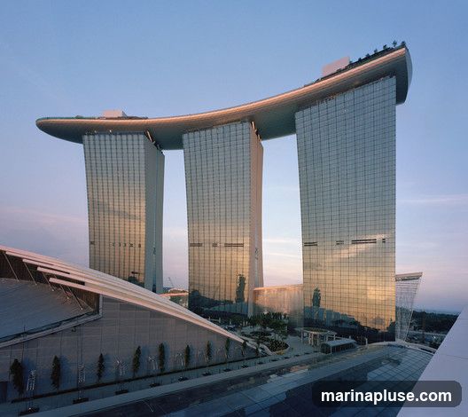 A wide shot of the Marina Bay Sands hotel in Singapore at dusk. The iconic building features three towers topped by a large, boat-shaped deck. The glass facades of the towers reflect the warm hues of the sunset. In the foreground, a modern, angular structure with a glass roof leads towards the hotel complex.