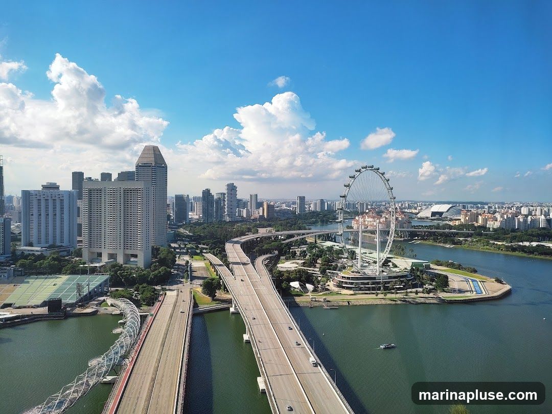 An aerial view of the Singapore skyline on a sunny day with a bright blue sky and scattered white clouds. The image features the Singapore Flyer Ferris wheel, several skyscrapers, elevated highways, and a body of water. A distinctive bridge with a spiral design is also visible.