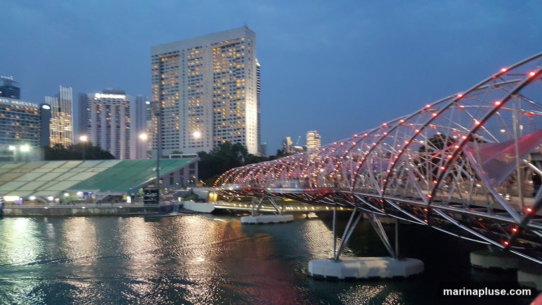 A wide shot of a city skyline at dusk, featuring several tall buildings illuminated with many lights. In the foreground, a modern, curved pedestrian bridge is visible, adorned with red lights along its structure. The bridge spans over a body of water that reflects the lights from the city and the bridge. The sky is a deep blue, indicating twilight.