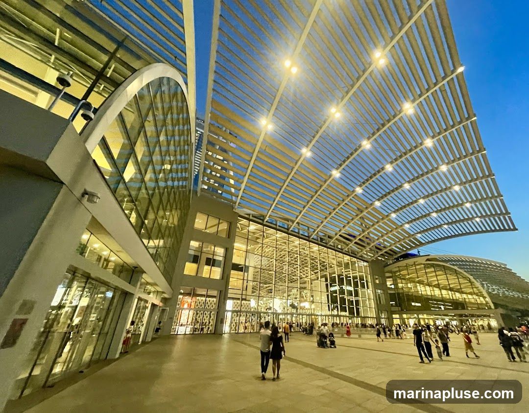 A wide-angle view of a modern shopping center's exterior at dusk. The architecture features a large, curved, slatted roof structure illuminated by numerous lights, extending over a wide plaza. The building itself has a striking facade with large glass windows and geometric architectural elements. People are seen walking in the plaza and entering/exiting the building. The sky is a deep blue, indicating twilight.