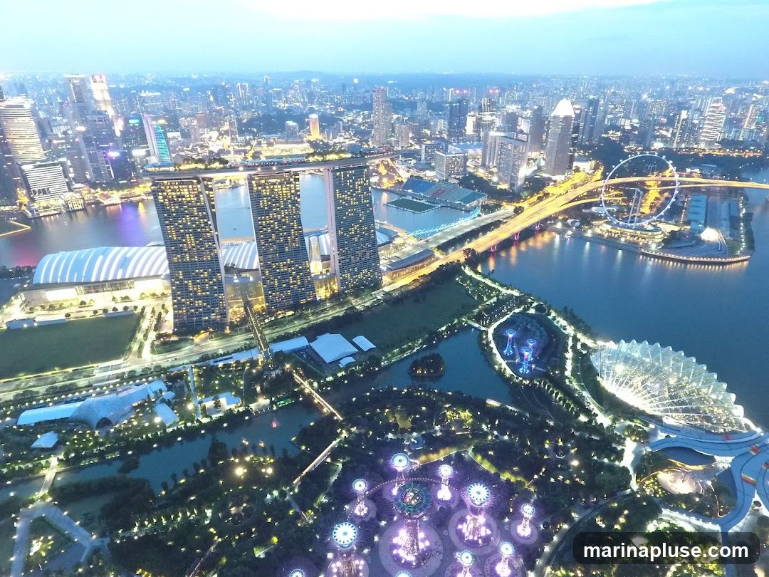 An aerial view of the Singapore skyline at dusk, showcasing the Marina Bay Sands hotel, Gardens by the Bay, and the Singapore Flyer. The city lights are beginning to illuminate the urban landscape, with the bay reflecting the glow of the buildings. The Gardens by the Bay features illuminated Supertrees and conservatories, creating a magical atmosphere.
