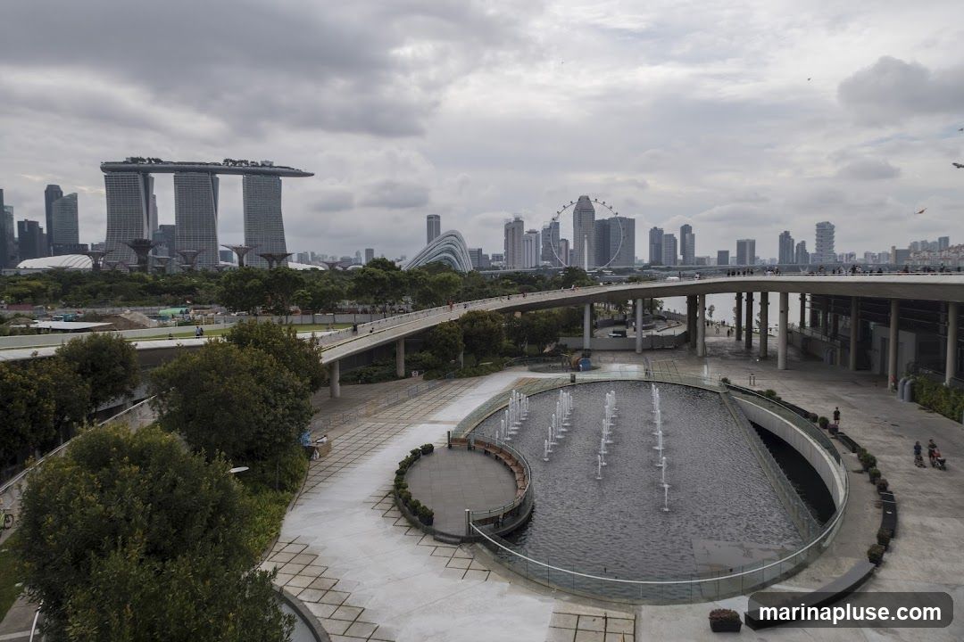 An aerial view of the Singapore skyline on a cloudy day, featuring the iconic Marina Bay Sands hotel, the Singapore Flyer Ferris wheel, and the Supertree Grove at Gardens by the Bay. In the foreground, a circular fountain with multiple jets is active in a paved plaza. A pedestrian walkway curves over the water, connecting different parts of the area. People are visible walking on the walkway and in the plaza.