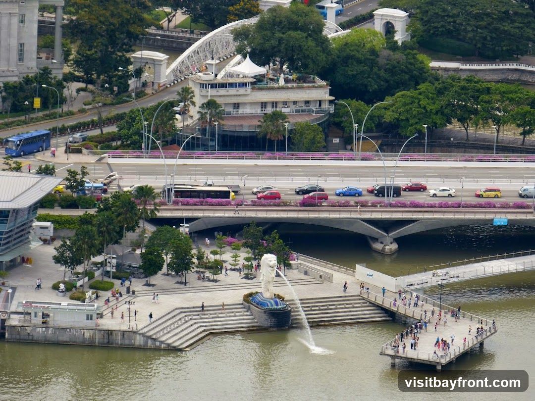 An aerial view of Merlion Park in Singapore, featuring the iconic Merlion statue spouting water into the bay. A pedestrian bridge extends from the park, and a busy road bridge with cars and buses crosses the water in the background. The cityscape includes modern architecture and lush greenery.