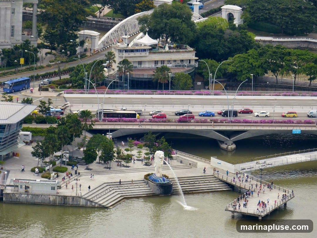 An aerial view of Merlion Park in Singapore, featuring the iconic Merlion statue spouting water into the bay. A pedestrian bridge extends from the park, and a busy road bridge with cars and buses crosses the water in the background. The cityscape includes modern architecture and lush greenery.