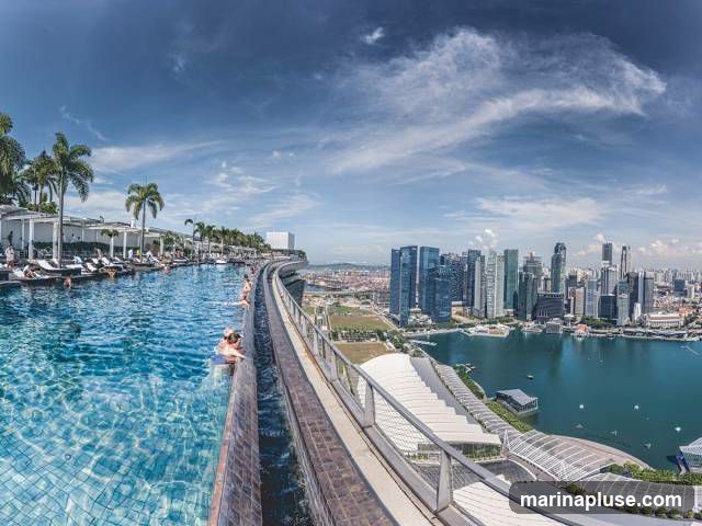 An expansive infinity pool with clear blue water is situated on a rooftop, offering a panoramic view of a bustling city skyline. Palm trees line the edge of the pool area, where several people are seen relaxing and swimming. The cityscape features numerous skyscrapers and a large body of water, under a bright blue sky with scattered clouds.