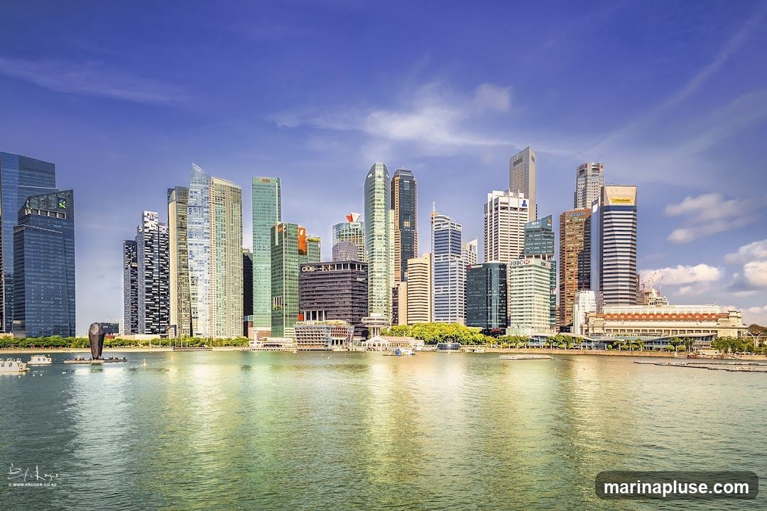 A panoramic view of the Singapore skyline with numerous skyscrapers of varying heights and architectural styles. The buildings are situated along the waterfront, with a body of water in the foreground reflecting the blue sky and clouds. Several boats are visible on the water. The image captures a bright, sunny day with a clear blue sky and scattered white clouds.