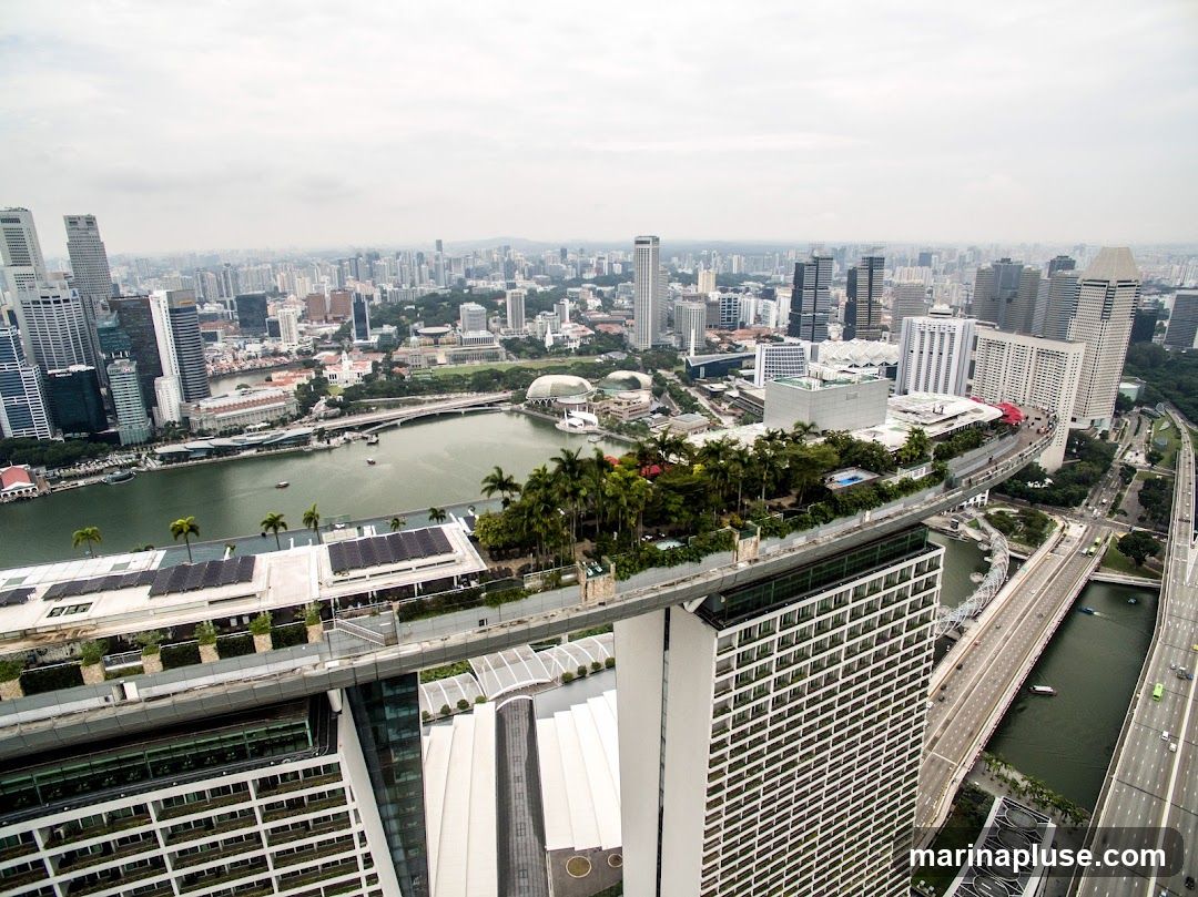 An aerial view of the Singapore skyline on an overcast day. The image features the Marina Bay Sands hotel with its iconic rooftop infinity pool and garden, as well as the surrounding cityscape with numerous skyscrapers, a river, and a highway.