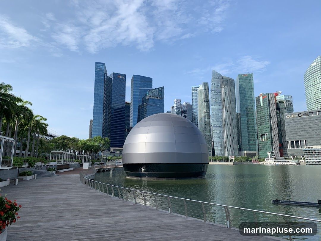 A wide shot of Marina Bay in Singapore on a clear day. In the foreground, a wooden boardwalk curves along the water's edge. To the left, palm trees and white structures line the promenade. In the center of the bay, a large, spherical, metallic building sits on the water. Behind it, the iconic skyscrapers of the Singapore skyline rise, including the DBS Bank building. The water reflects the blue sky and the surrounding architecture.