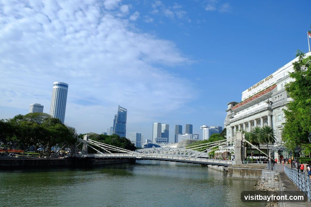 A wide shot of the Singapore River on a clear day. A suspension bridge spans the river, with modern skyscrapers and the historic Fullerton Hotel visible in the background. Lush green trees line the riverbanks, and a promenade with people walking can be seen on the right.