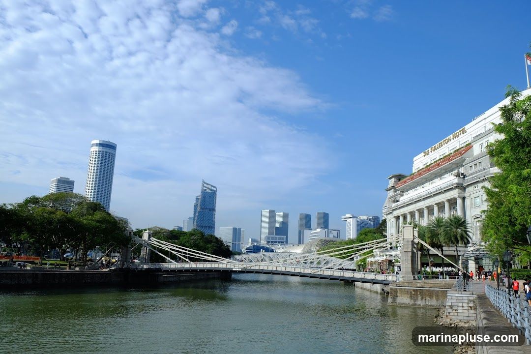 A wide shot of the Singapore River on a clear day. A suspension bridge spans the river, with modern skyscrapers and the historic Fullerton Hotel visible in the background. Lush green trees line the riverbanks, and a promenade with people walking can be seen on the right.