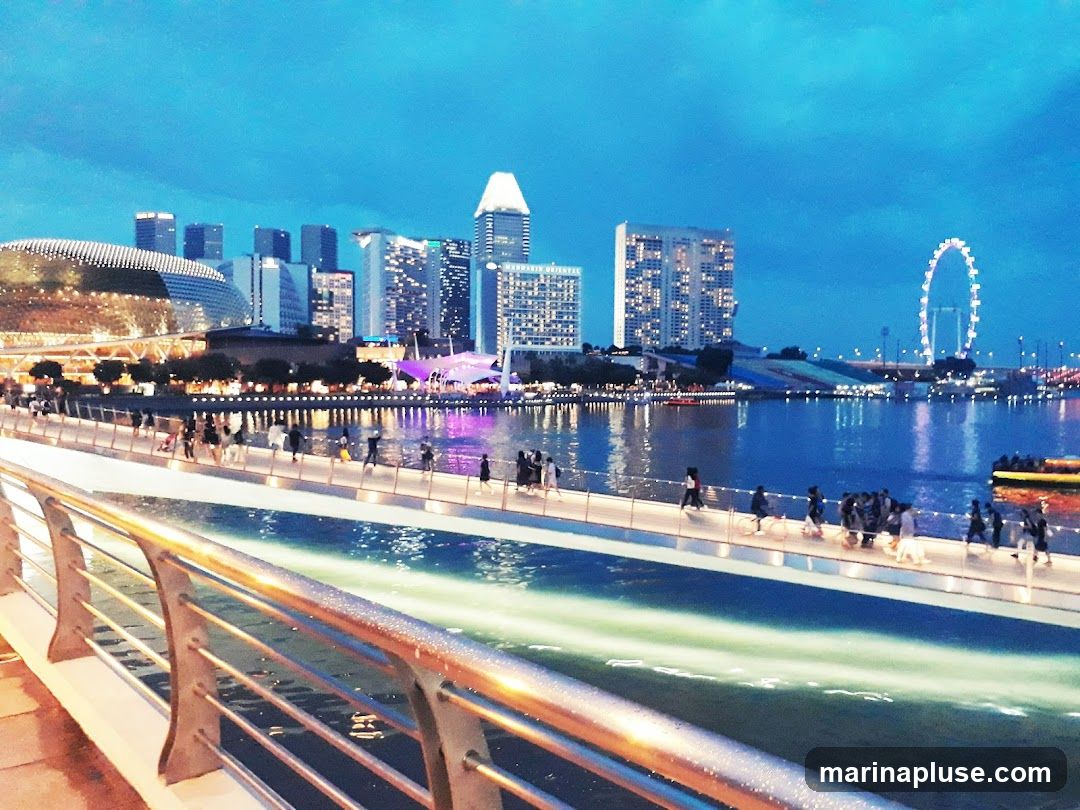A wide shot of a city skyline at dusk, with a body of water in the foreground. A pedestrian bridge spans across the water, with many people walking on it. The buildings in the background are illuminated, and a Ferris wheel is visible in the distance. The sky is a deep blue, transitioning from day to night.