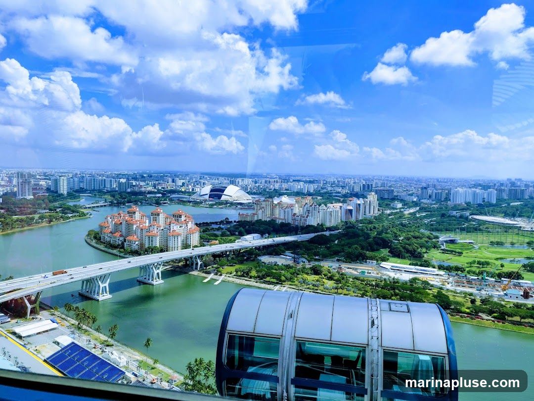 A wide-angle view of a sprawling cityscape with a river winding through it. In the foreground, a segment of a large, modern observation wheel capsule is visible. The capsule has large windows offering a clear view of the scenery. The city features numerous buildings of varying heights, including residential complexes and a distinctive stadium with a domed roof in the distance. A prominent bridge spans the river, with traffic visible on it. Lush green trees and parks are interspersed throughout the urban landscape. The sky is a vibrant blue, dotted with fluffy white clouds.