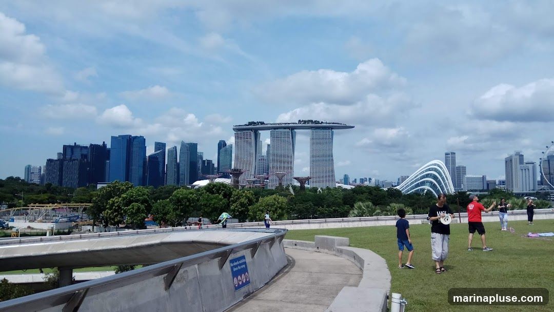 A panoramic view of the Singapore skyline, featuring the iconic Marina Bay Sands hotel, the Supertrees at Gardens by the Bay, and other modern skyscrapers. In the foreground, a grassy area with a concrete walkway, where several people are seen enjoying the day, some flying kites. The sky is partly cloudy.