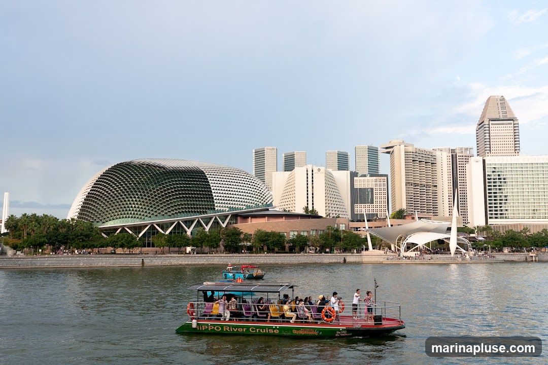 A wide shot of a river with several boats on it, in front of a city skyline. The most prominent building in the skyline is a large, dome-shaped structure with a distinctive patterned facade. Other buildings include modern skyscrapers and a white, tent-like structure. The boat in the foreground is a tour boat with passengers visible. The water is calm and reflects the sky.