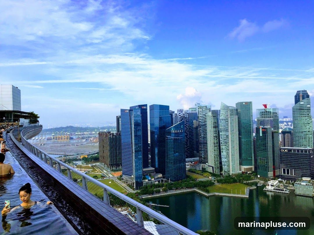 A woman takes a selfie in an infinity pool on a rooftop, with a panoramic view of the Singapore skyline and a busy port in the background. The sky is blue with scattered clouds.