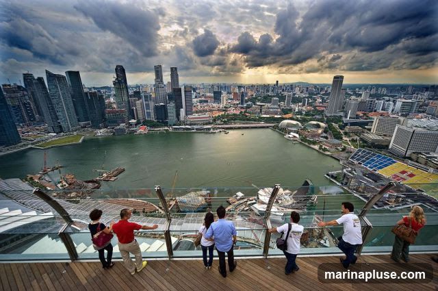 A group of tourists stand on a wooden deck with a glass railing, admiring a sprawling cityscape and a large body of water. The city features numerous skyscrapers and modern architecture. The sky is dramatic with heavy, dark clouds interspersed with rays of sunlight.