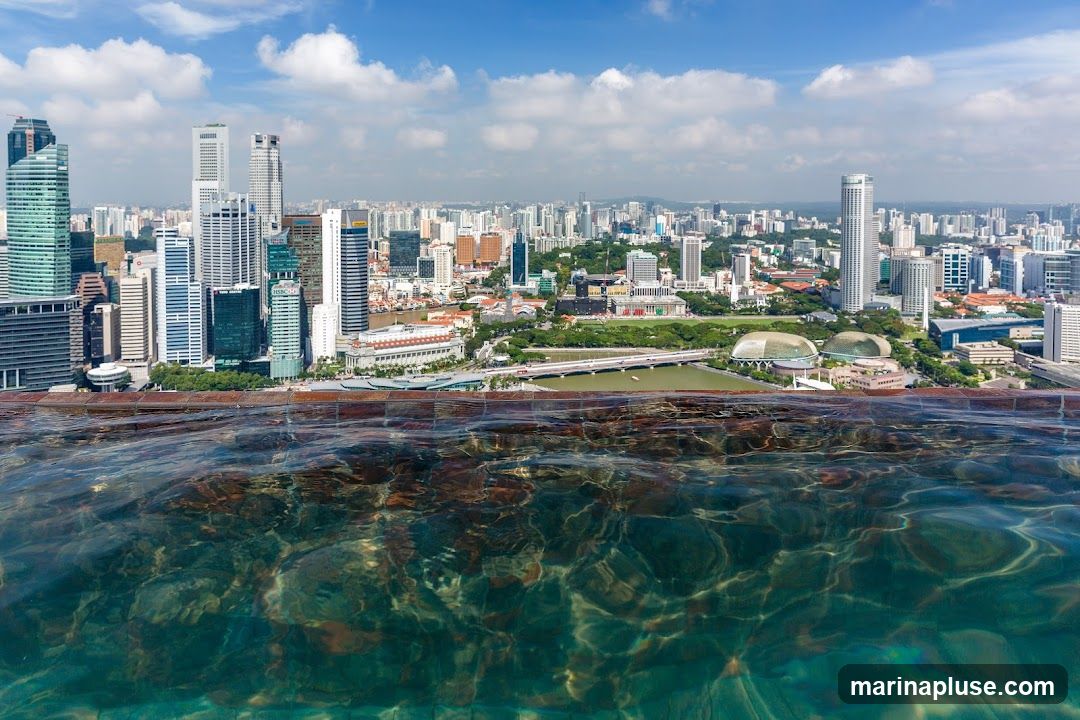An infinity pool with clear blue water in the foreground, reflecting sunlight and the sky. Beyond the pool, a panoramic view of the Singapore skyline unfolds, featuring numerous modern skyscrapers of varying heights and architectural styles. The cityscape extends to the horizon under a bright blue sky dotted with white clouds. In the mid-ground, a body of water with a bridge and some boats can be seen, along with green park areas and distinctive domed buildings.