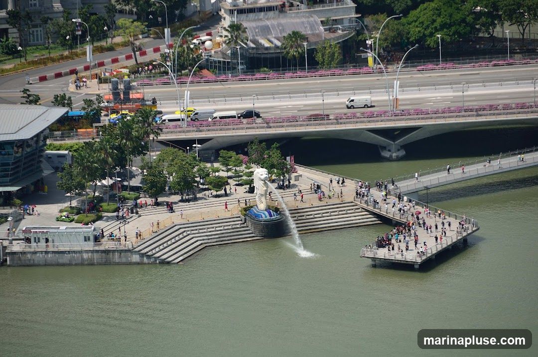 An aerial view of the Merlion fountain in Singapore, with the Marina Bay Sands hotel and a bridge in the background. Many people are gathered around the fountain and on the surrounding walkways.