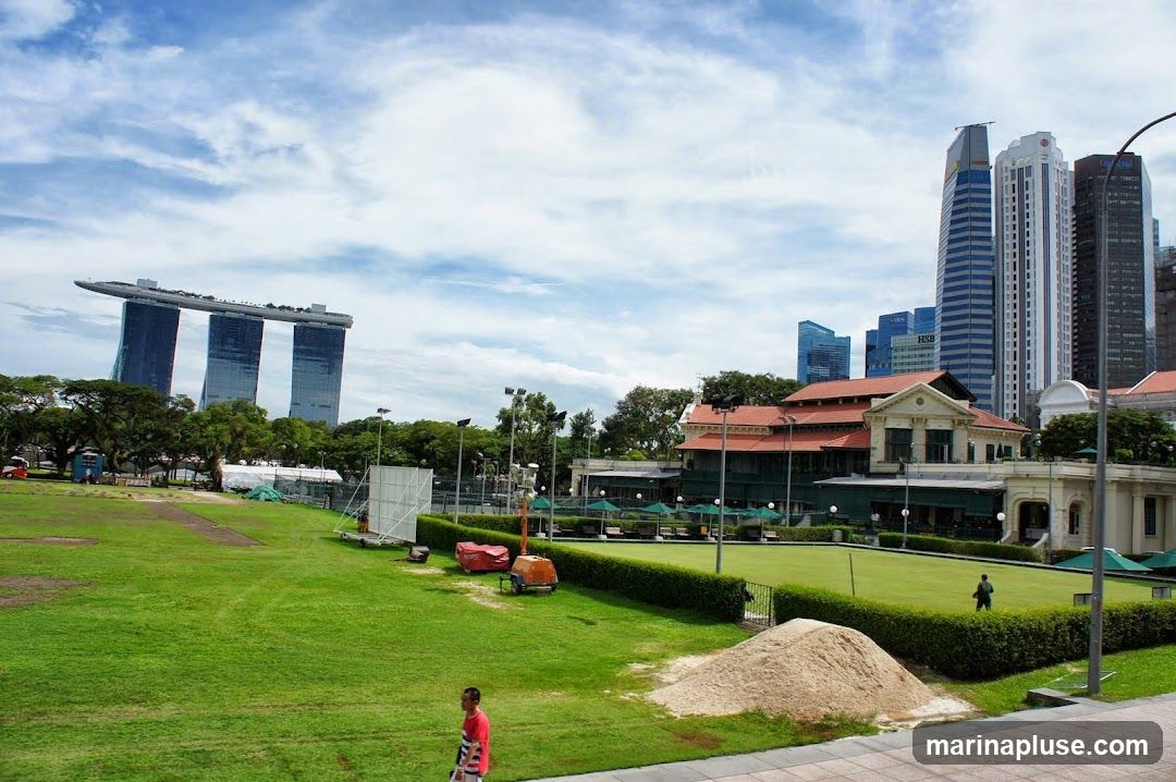 A wide shot of the Singapore skyline featuring the iconic Marina Bay Sands hotel in the distance. In the foreground, a lush green lawn is visible, with a building that appears to be a club or restaurant with outdoor seating. Several tall skyscrapers form the backdrop of the city. The sky is partly cloudy.