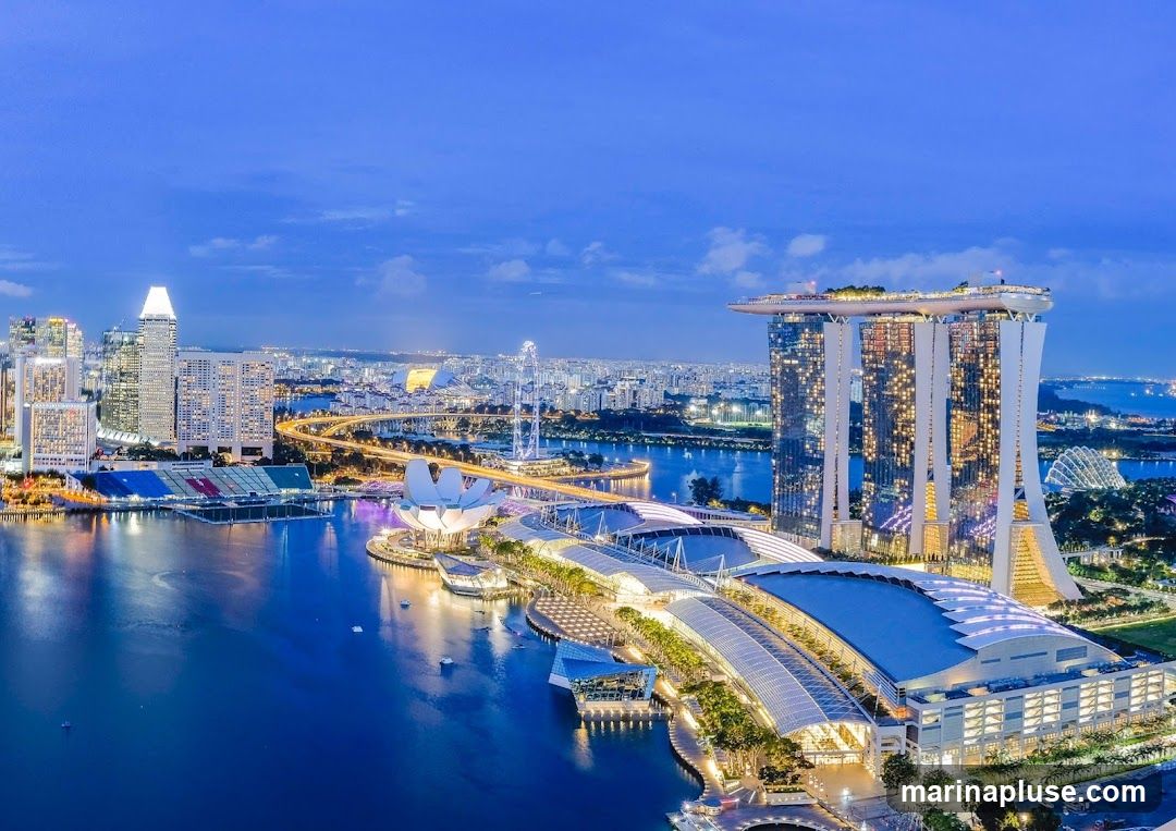 An aerial view of the Singapore skyline at dusk, featuring the Marina Bay Sands hotel, the ArtScience Museum, and other prominent buildings. The city lights are illuminated, reflecting on the water.