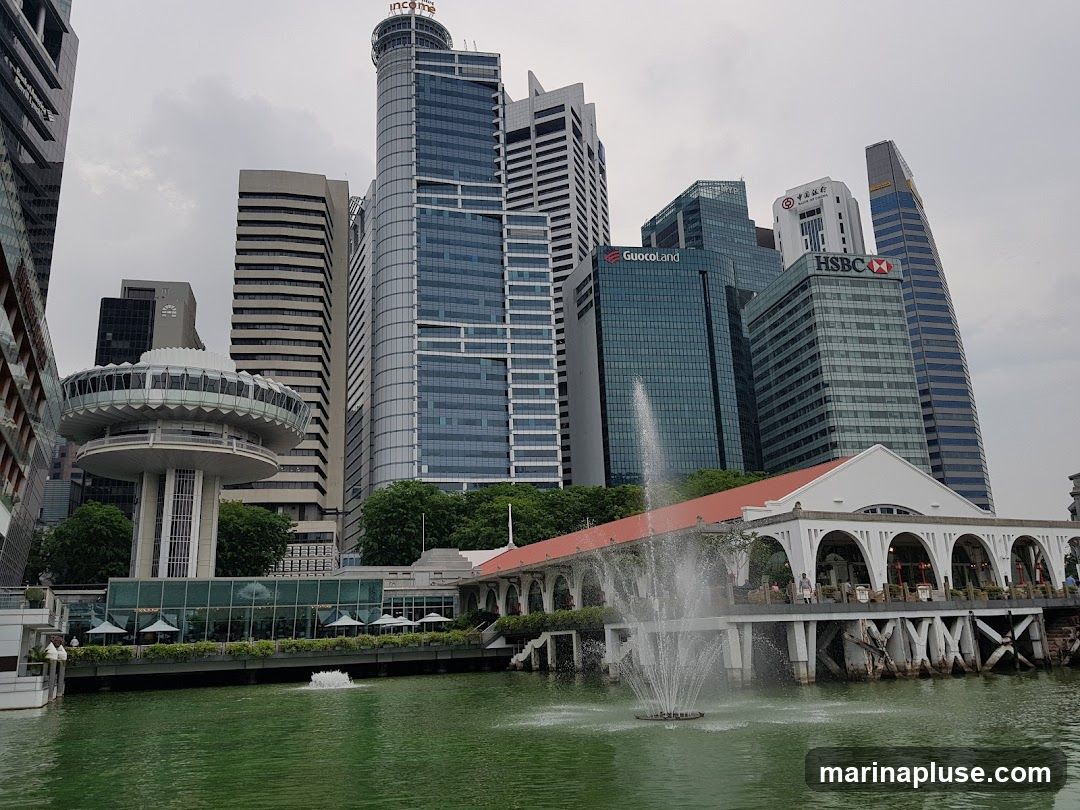 A wide shot of a city skyline featuring numerous modern skyscrapers and a distinctive white building with arched walkways situated along a body of green water. A fountain sprays water in the foreground. The sky is overcast.