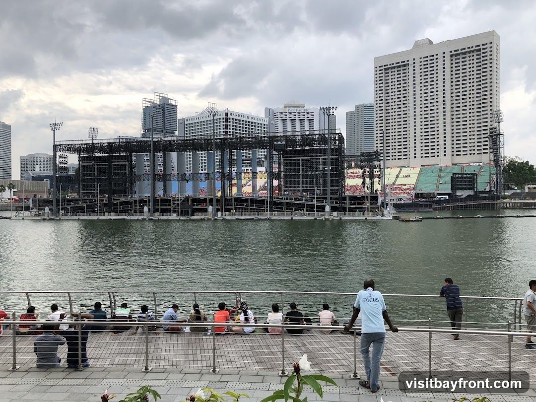 A group of people are seated on a wooden boardwalk, facing a large outdoor stage structure across a body of water. The stage appears to be under construction or set up for an event, with scaffolding and screens visible. In the background, several tall buildings form a city skyline under a cloudy sky.